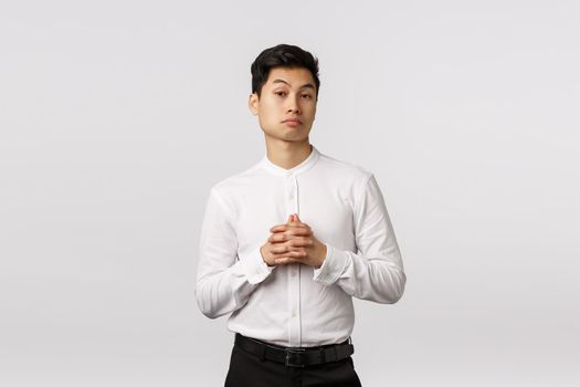 Skeptical and impressed young asian businessman consider something, clench hands together, raising eyebrows pleased, listen interesting offer, have business meeting, standing white background.