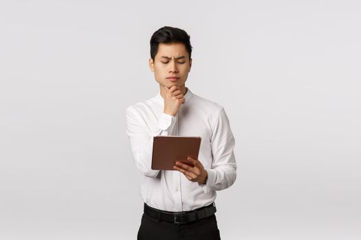 Pensive attractive asian businessman making hard choice, looking at digital tablet and thinking what order girlfriend online, reading documents on device screen, frowning thoughtful, white background.