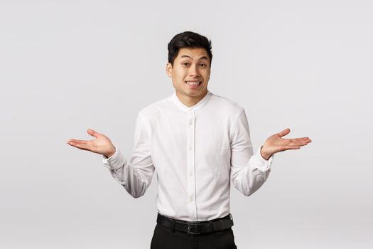 Oops sorry but cant help. Indecisive cute asian young man, businessman dont know how situation happened, explain himself, shrugging and smiling awkward, standing unsure white background.