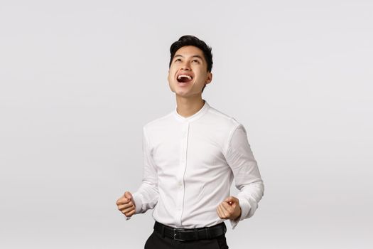 Yeah baby, thanks god I won. Man celebrating successful deal. Pleased, rejoicing attractive young asian guy fist pump, say yes at sky and smiling relieved, achieve goal, winning, white background.