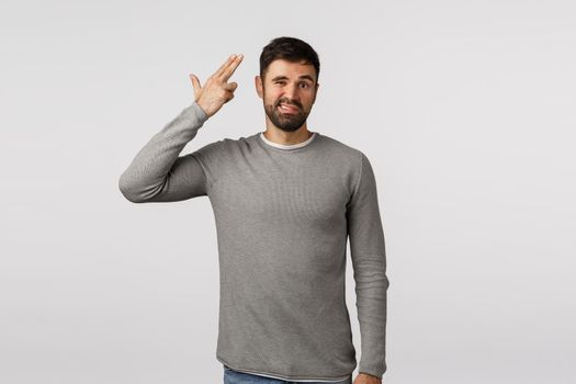 Kill me please. Bothered and tired, annoyed attractive caucasian bearded man in grey sweater, shooting from finger gun at head and grimacing, standing irritated, hear nonsense, white background.