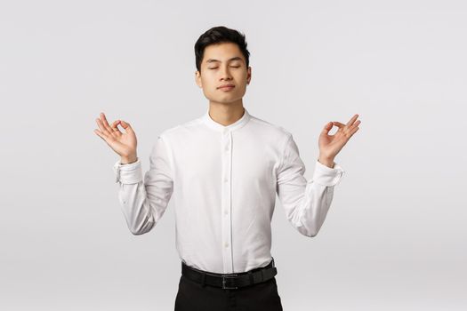 Peaceful and determined attractive asian businessman, calm down, release stress, close eyes and meditating in office, breathing practice with hands sideways, mudra or zen gesture, do yoga.