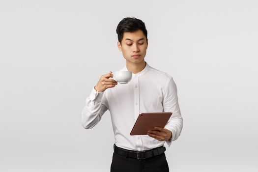 Business, technology and finance concept. Serious-looking elegant and stylish, successful male entrepreneur reading news in digital tablet, drinking coffee from cup, studying documents online.
