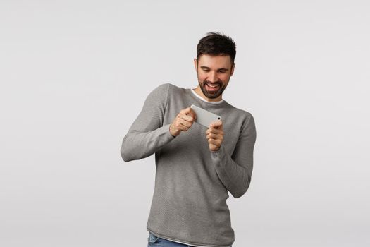 One more level. Cheerful good-looking bearded adult male in grey sweater, holding smartphone horizontally and bending to pass racing circle, playing application, mobile game, smiling joyfully.