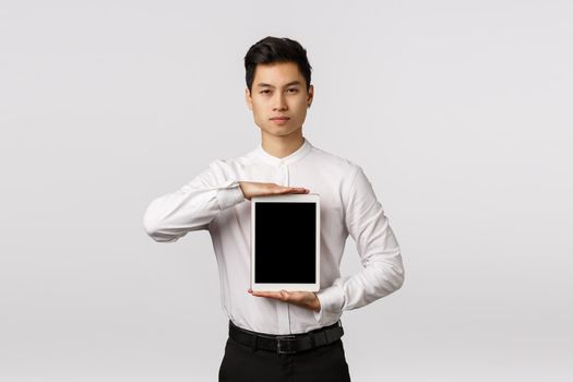 Guy knows exactly what you need. Serious-looking sassy and elegant asian man in formal outfit, holding digital tablet over chest, showing device screen, promote application or corporate link.