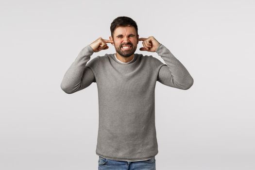 Distressed and disturbed guy bothered by loud neighbour, grimacing, clench teeth and squinting, shut ears, plug fingers unwilling hear awful sound, telling turn-off music, standing white background.