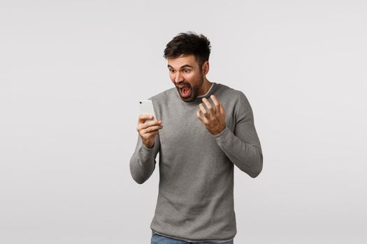 Outraged and furious pissed, tensed bearded man in grey sweater, holding smartphone, cursing, swearing at mobile display, yelling phone screen and shaking hands, standing white background.