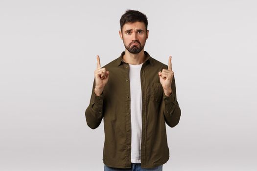 Sad and distressed lonely, miserable handsome bearded male in coat, pulling grimace with jealous, regret face, frowning pointing fingers up, telling disappointing bad news, complain, white background.