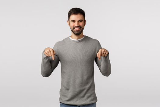 Time to see it, look here. Happy, charismatic friendly-looking smiling bearded male in grey sweater, invite event, share link, discuss promo offer, pointing down to promote product, white background.