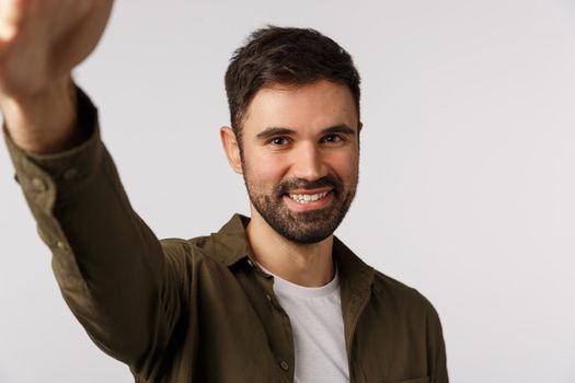 Handsome cheerful bearded man in coat want upload new photo gay dating application, hold smartphone with arm and smiling camera, taking selfie, standing white background joyful. Copy space