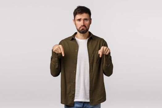 Man having disappointing bad news. Upset and miserable, unhappy young bearded man feeling regret or jealous, pulling sad grimace, frowning, pointing down complaining, white background.