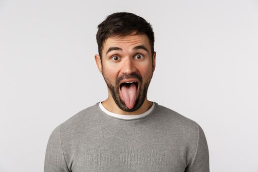 Carefree funny and playful, goofy guy with beard, wear grey sweater, showing tongue, open mouth and stare camera, fool around, showing inner self, go wild and relieved, standing white background.