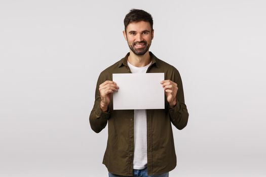 Assertive smiling cheerful bearded caucasian man in coat, holding blank paper near chest and grinning, searching for employees, have job or produce offer, place corporate banner, white background.