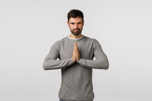 Determined and motivated good-looking bearded caucasian guy in grey sweater, bowing politely with hands pressed together in namaste, asian greeting, smiling, praying over white background.