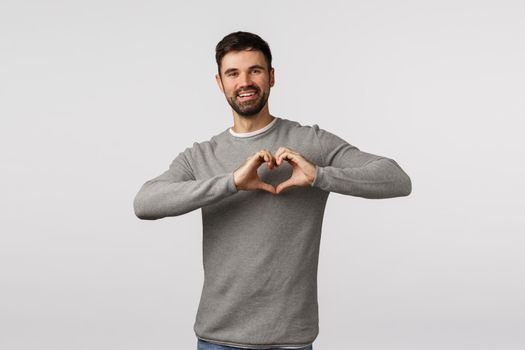 Love, family and care concept. Charming bearded boyfriend express affection and tender feelings, make heart sign over chest and smiling, cherish relationship, adore partner, white background.
