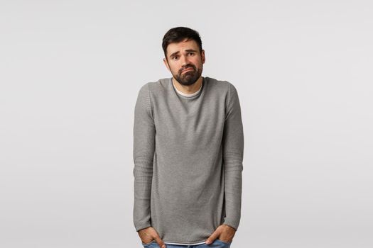 Modest, unconfident young man feeling loser, frustrated and sad standing in grey sweater over white background, shrugging holding hands in pockets, smirk and frowning distressed, indecisive.