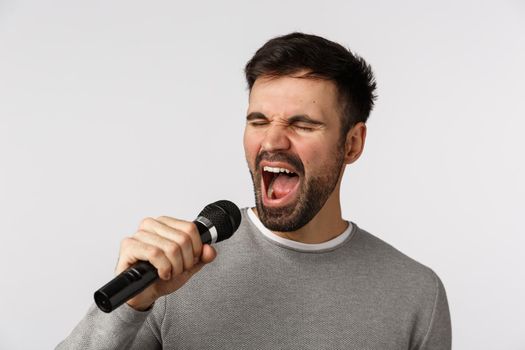 Close-up shot carefree cheerful gay man with beard, grey sweater, singing along upbeat song, holding microphone, carried away with lyrics, attend karaoke party night, standing white background.