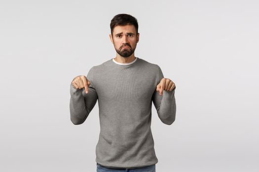 Gloomy and depressed, upset sulky boyfriend with beard in grey sweater, pointing down, pulling smile down distressed and jealous, feel regret or disappointment, standing white background.
