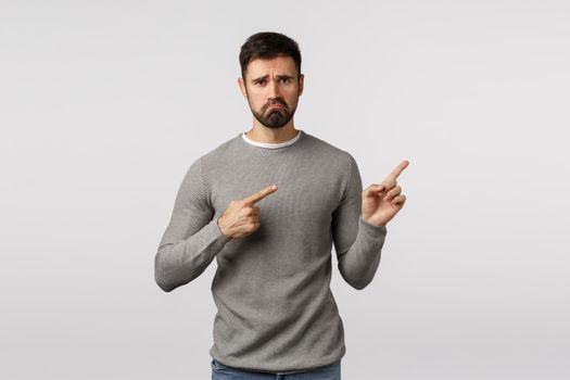 Gloomy, depressed and lonely miserable handsome bearded guy in grey sweater, pointing upper right corner, pulling grumpy, sulking face, frowning complain, feel jealous or regret, showing bad news.