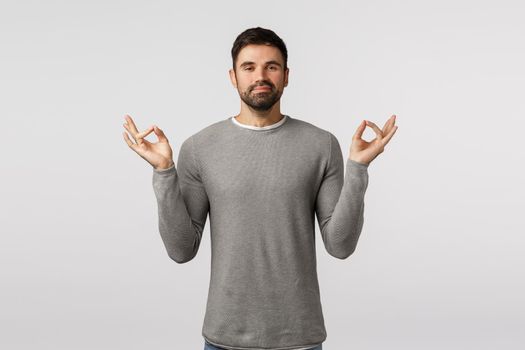 Peaceful and cheerful, smiling happy bearded male in grey sweater feeling okay after meditation, relief stress, hold hands sideways mudra, zen gesture, reach nirvana, practice yoga, white background.