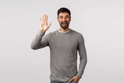 Friendly, cheerful and outgoing smiling bearded caucasian man, greeting neighbour raising arm and waving in hello, hi gesture, see person as waiting in meeting spot, standing white background.