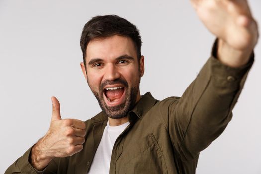 Excited, upbeat handsome bearded man absolutely adore party, like event, hold smartphone with one arm and show thumb-up, taking selfie, record vlog, online stream, standing white background.