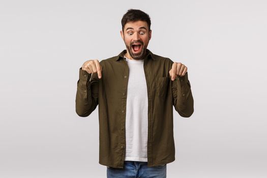 Excited, happy and cheerful handsome bearded man in coat, pointing down and looking bottom advertisement with fascinated, overjoy expression as see wonderful christmas promo, white background.