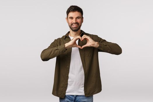 Valentines day, affection and relationship concept. Cheerful tender bearded male in coat, showing heart gesture near chest, express love or adornment, smiling cute, express gratitude or affection.