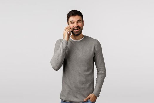 Carefree enthusiastic, bearded male in grey sweater talking on phone with pleased, happy smile, hold hand in pockets, use smartphone calling via internet messanger, standing white background.