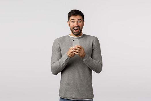 Excited, cheerful good-looking caucasian male in grey sweater, holding smartphone, smiling fascinated and thrilled, photographing interesting lecture, standing wondered and upbeat, white background.