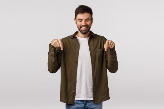 Satisfied cute bearded male found good place hang out, checking out nice offer and smiling, considering buy it, make choice, pointing down and grinning satisfied, standing white background.