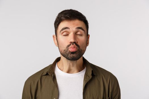 Funny and cute handsome bearded guy in coat, close eyes and folding lips as awaiting for passionate kiss, sending mwah, having romantic date on valentines day, standing white background.