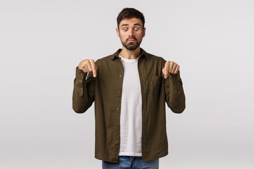 Alarmed man standing in stupor over white background seeing poisoned spider crawrling his foot, stare scared and anxious, pointing down, not moving and breathing frightened, white background.