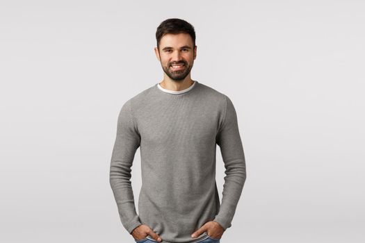 Cheerful, charismatic bearded, masculine guy in grey sweater, hold hands pockets, carefree pose, smiling delighted, speaking to client, discuss business, satisfied with good results, white background.