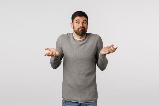 Funny bearded male in grey sweater, shrugging with hands raised sideways, smirk and look away as if dont know, cant help, standing indecisive, hiding truth, unwilling say, posing white background.