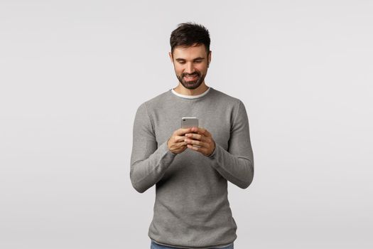 Handsome bearded caucasian guy in grey sweater making playlist, holding smartphone and listen music wireless headphones, enjoy songs using headphones, watching video in crowded place, smiling.