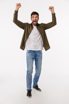 Success, achievement and victory concept. Vertical full-length shot glad and happy triumphing young bearded man in jeans and coat, raise hands up in hooray, win gesture, celebrating great news.