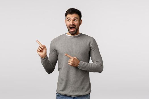 Excited, surprised very lucky handsome bearded boyfriend in grey sweater drop jaw from thrill and astonishment, smiling amused, check out awesome promo, hot sale, standing white background overjoy.
