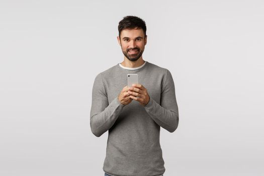 Handsome smiling and happy caucasian bearded man taking photo on smartphone, holding mobile phone near chest, grinning, photographing, record video, stream concert online, white background.
