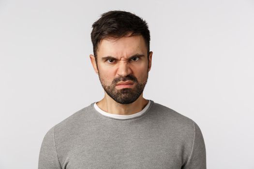 Close-up studio shot hateful and outraged, aggressive scary bearded adult man in grey sweater, grimacing, wrinkle nose and look with disgrace, disdain or scorn, standing bothered white background.