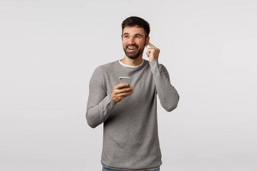 Cheerful good-looking, amazed caucasian guy enjoy awesome sound quality of headphones, put wireless earphones, increase volume, holding smartphone, looking away dreamy and smiling, white background.