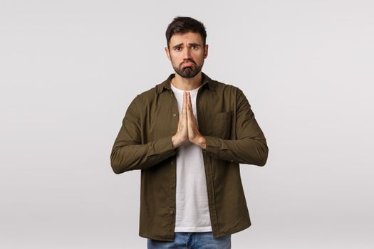Upset, miserable and gloomy cute bearded guy in casual outfit, begging for help need something, press palms together asking offer, praying, supplicating, ask apology, white background.