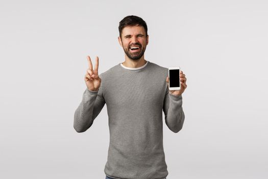 Funny and emotive, charismatic bearded man holding smartphone, make peace, victory sign, showing mobile screen as advertise product, shopping site or tracking order application, white background.
