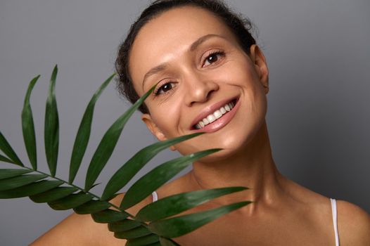 Attractive woman with beautiful smile and clean skin looks at the camera holding a palm leaf isolated over gray background with copy space. Spa, beauty treatment, anti-aging, natural cosmetics concept