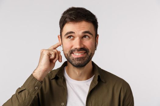 Carefree smiling handsome bearded adult male in coat, looking up dreamy and joyful, touching wireless earphone to tune in, change volume or finish call, standing white background happy.