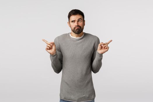 Indecisive and clueless handsome bearded guy in grey sweater, shrugging troubled make choice, have two variants, choosing between products in shopping market, grimacing unsure, pointing sideways.