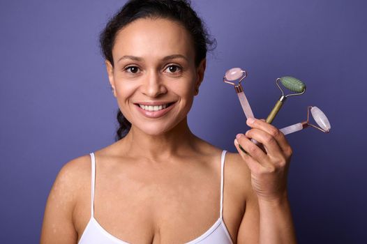 Beauty portrait of a young African pretty woman with vitiligo skin problems, in white underwear smiles with a beautiful toothy smile holding colorful facial roller massagers near her face.