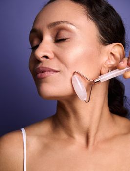 Close-up. Middle aged African American pretty woman doing smoothing anti-aging face massage, massaging her cheek with pink jade stone roller massager, isolated on purple background with copy ad space