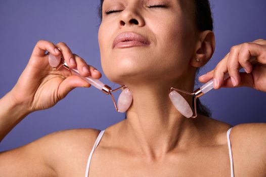 Macro beauty portrait of a relaxed beautiful woman massaging her neck with jade roller massagers, isolated over purple background with copy space for ads. Face lift anti-aging treatment concept