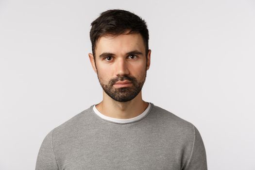 Close-up shot serious and determined handsome, adult masculine bearded man in grey sweater, looking with casual expression camera, standing still, awaiting important news, white background.